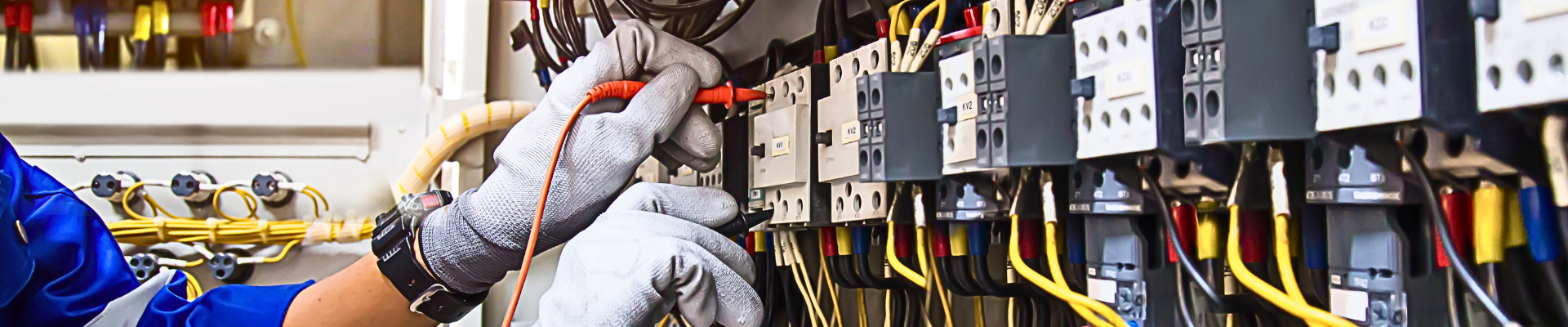 A person wearing gloves uses a tool on a complex electrical panel with numerous wires and components.