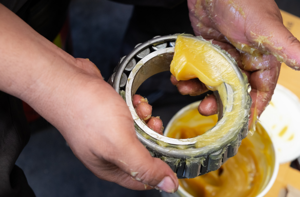 A person applies yellow grease to a large metal wheel bearing using their hands, with a container of the grease visible nearby.
