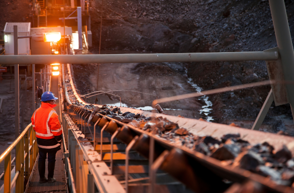 A person in a safety vest and helmet observes an outdoor mining conveyor belt transporting raw materials. The scene is lit by industrial lighting, highlighting the machinery and rocky terrain. The sky is dim, suggesting early morning or late evening.