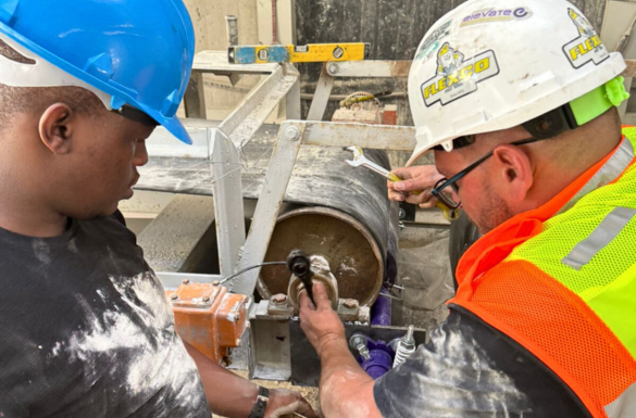 Two men wearing hard hats observe as a conveyor belt trainer is installed.