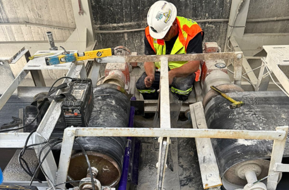 A worker wearing a white helmet and high-visibility vest is seated on industrial equipment inside a factory setting. The equipment includes large metal cylinders, wiring, and various tools.